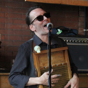 Photo of Josh Broadbent, singing into a microphone, while wearing dark sunglasses and playing a washboard