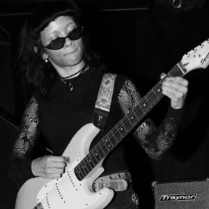Photo of Sophia Edross, wearing sunglasses and playing an electric guitar